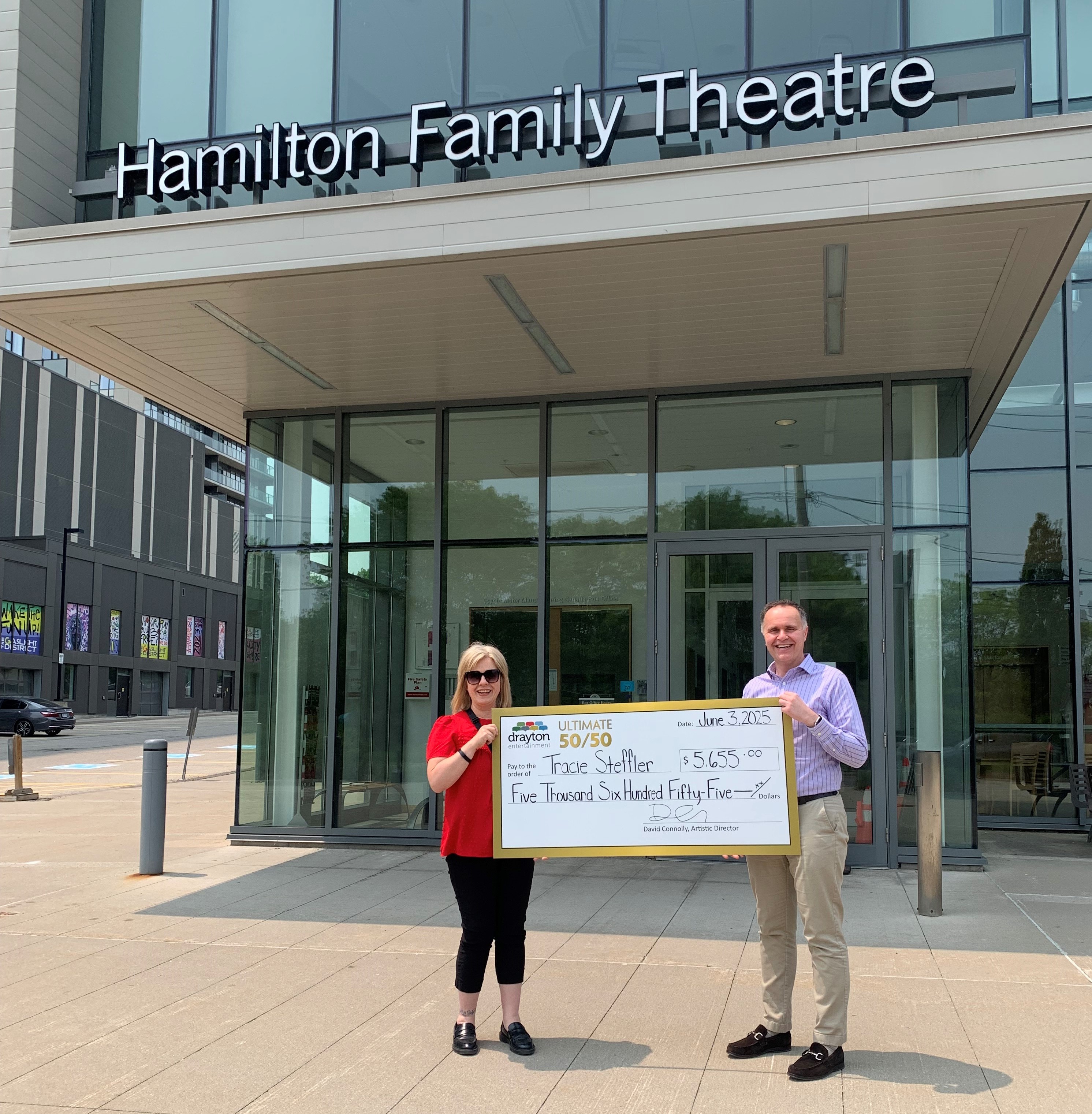 Tracie Settler (left) stands outside the Hamilton Family Theatre Cambridge holding a large presentation cheque for $5,655 alongside Drayton Entertainment Artistic Director David Connolly (right). The cheque, dated June 3, 2025, recognizes Tracie as the May 2025 winner of the Ultimate 50/50 draw.